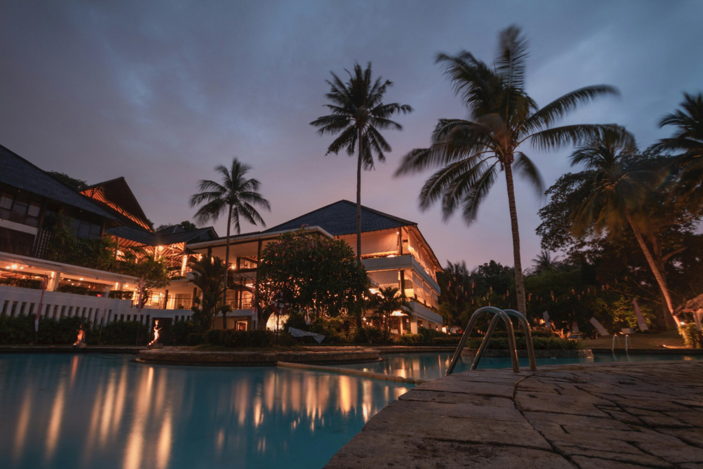 A luxurious mansion with glowing lights at dusk, featuring a serene swimming pool, tall palm trees, and lush tropical landscaping
